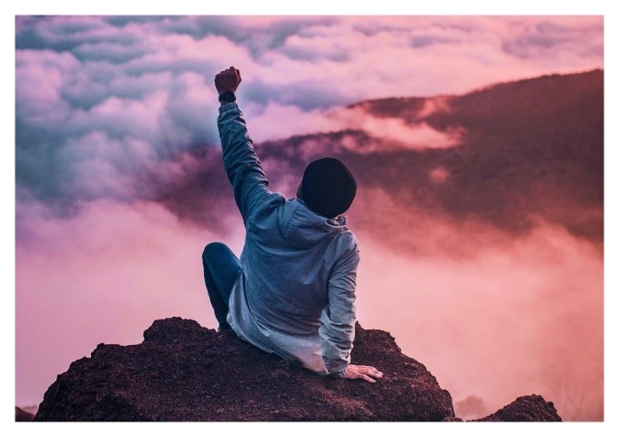 Persona sentada en la cima de una montaña, con la mano hacia arriba y el puño cerrado.